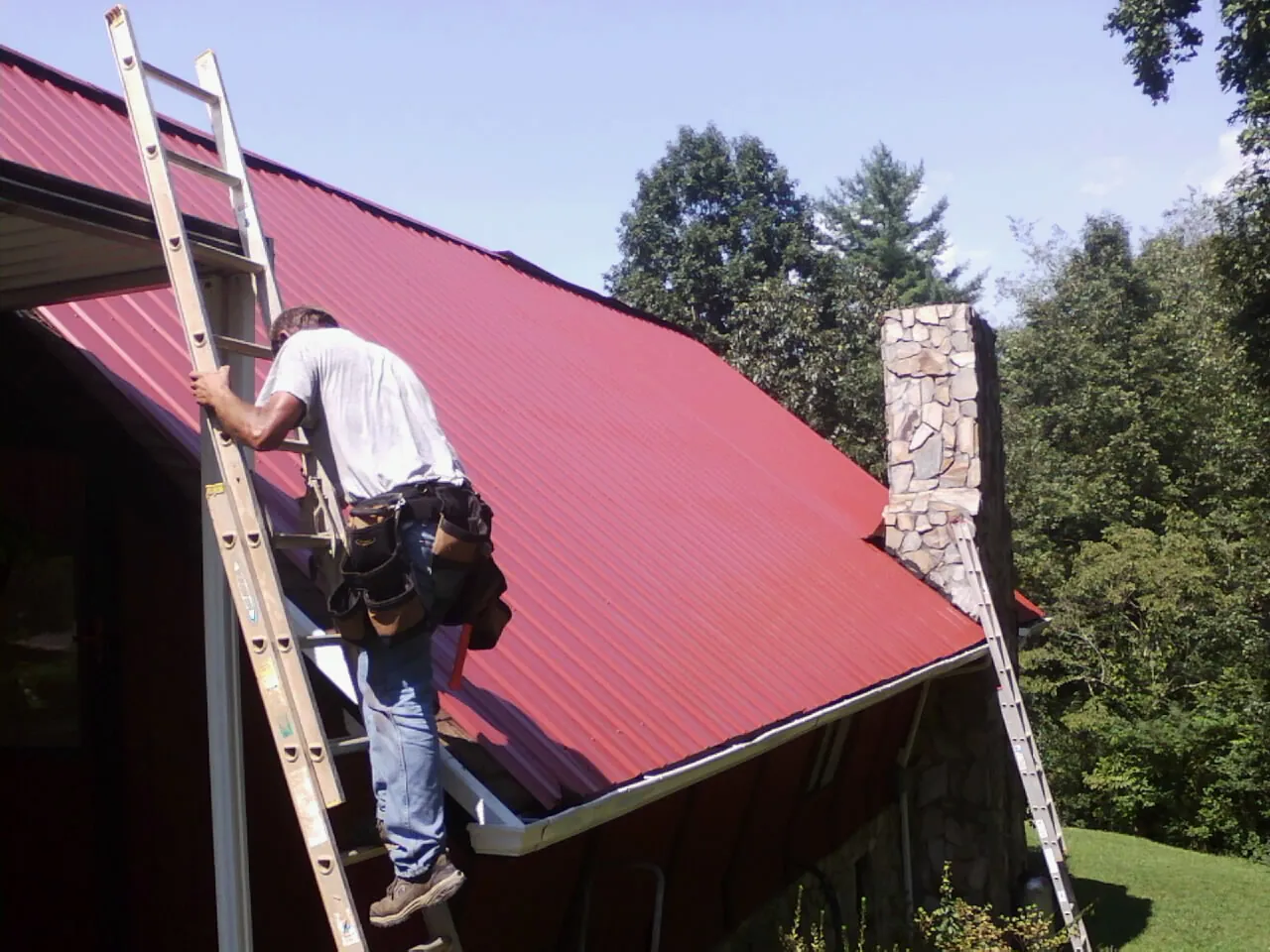 Roofer on a ladder beside a red metal roof and stone chimney — Soffit & Fascia Repair by Roofing Brothers in Havre De Grace